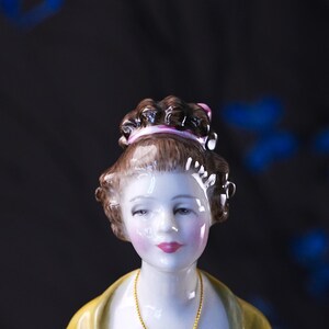 May include: A porcelain figurine of a person with brown hair styled in an updo with a pink ribbon. The figure has a pale complexion with rosy cheeks, red lips, and a yellow garment. A thin gold-colored chain hangs around the neck of the figure.