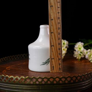 K&ouml;nnte beinhalten: Eine kleine, wei&szlig;e Keramikvase mit schmalem Hals und rundem K&ouml;rper. Die Vase hat ein gr&uuml;nes Blattdesign und Text auf der Seite. Ein Lineal zeigt, dass die Vase etwa 10 cm hoch ist. Die Vase steht auf einem dekorativen Tablett.