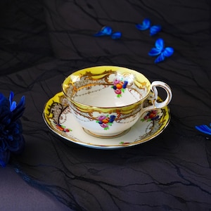 Peut inclure: Un ensemble tasse et soucoupe en porcelaine avec un bord jaune vif, des volutes dor&eacute;es complexes et des grappes de fruits et de fleurs color&eacute;es peintes &agrave; la main. La tasse poss&egrave;de une anse incurv&eacute;e et repose sur la soucoupe assortie.