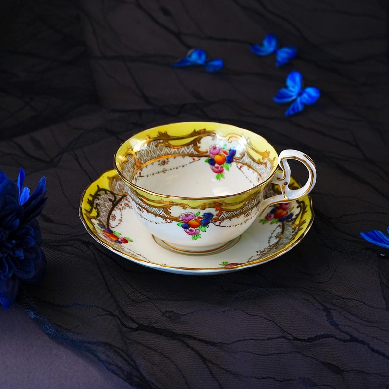 Peut inclure: Un ensemble tasse et soucoupe en porcelaine avec un bord jaune vif, des volutes dor&eacute;es complexes et des grappes de fruits et de fleurs color&eacute;es peintes &agrave; la main. La tasse poss&egrave;de une anse incurv&eacute;e et repose sur la soucoupe assortie.