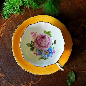 Peut inclure: Un service à thé vintage composé d'une tasse et d'une soucoupe. La tasse présente une rose rose et un motif floral bleu sur fond blanc. La soucoupe et le bord de la tasse sont jaune vif avec une bordure dorée. L'ensemble est posé sur une surface en bois foncé.