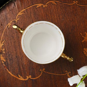 Puede incluir: Una taza de t&eacute; de porcelana blanca con dos asas doradas. El borde interior presenta una inscripci&oacute;n dorada que dice: TO CELEBRATE THE GOLDEN JUBILEE OF HER MAJESTY QUEEN ELIZABETH II. La taza tiene un interior circular liso y un borde dorado.