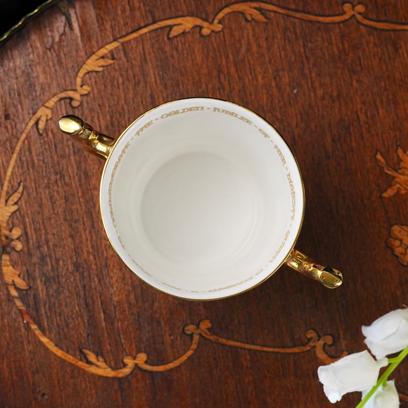 Puede incluir: Una taza de t&eacute; de porcelana blanca con dos asas doradas. El borde interior presenta una inscripci&oacute;n dorada que dice: TO CELEBRATE THE GOLDEN JUBILEE OF HER MAJESTY QUEEN ELIZABETH II. La taza tiene un interior circular liso y un borde dorado.