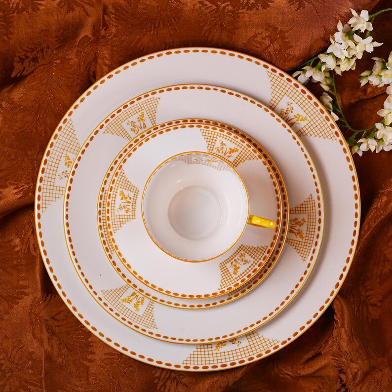 Op de afbeelding: Een witte theekop en schotelset met een gele handgreep, geplaatst op een stapel witte borden. De borden en de kop hebben een gouden rand en een goud-geel bloemmotief. De set is geplaatst op een bruin tafelkleed.