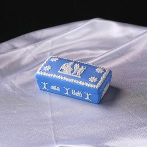 May include: A rectangular blue trinket box with white relief detailing. The box features floral and classical figures in relief. The edges are trimmed with a white floral pattern. The box is set on a white satin surface.