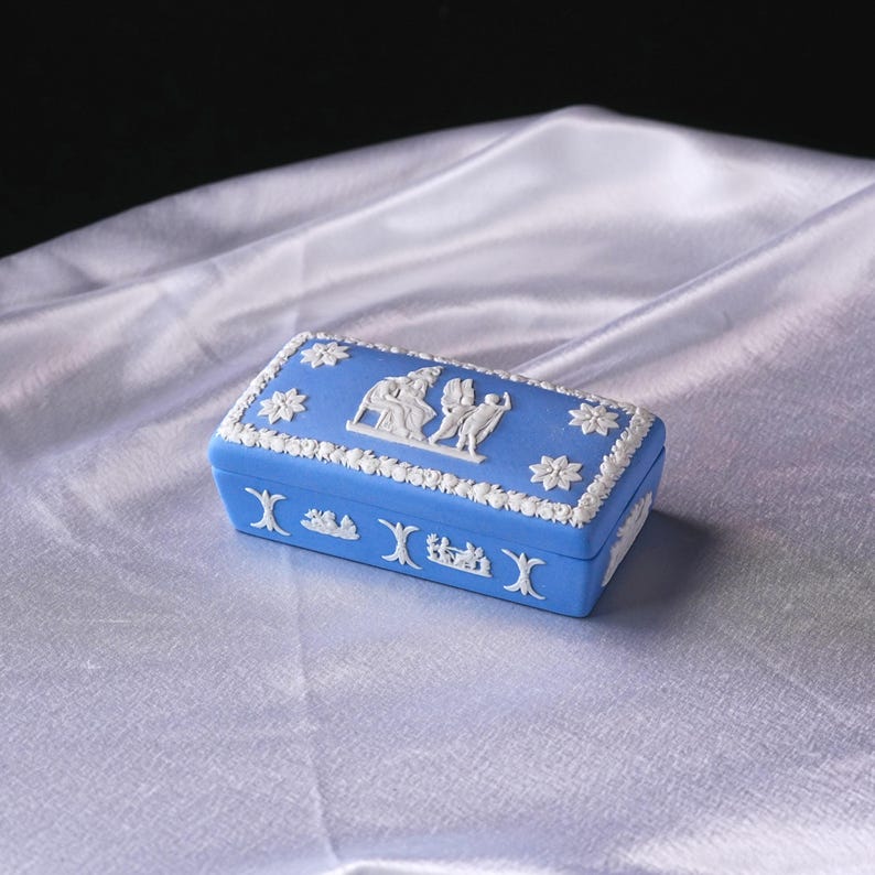 May include: A rectangular blue trinket box with white relief detailing. The box features floral and classical figures in relief. The edges are trimmed with a white floral pattern. The box is set on a white satin surface.