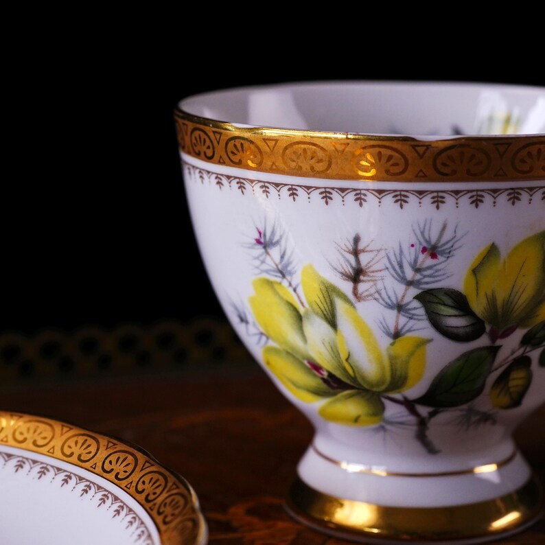 Peut inclure: Tasse blanche avec un bord et un pied dor&eacute;s, orn&eacute;e d'un motif floral jaune avec des feuilles vertes et des accents gris. La tasse a un piedestal et une soucoupe assortie est partiellement visible.