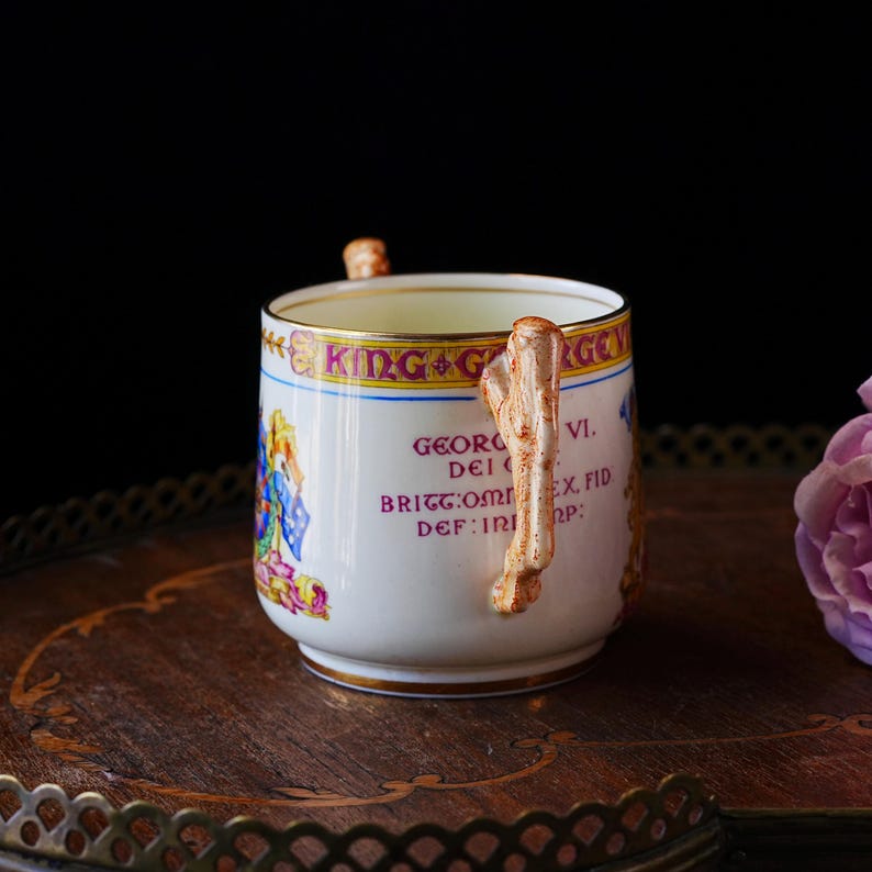 Puede incluir: Una taza de t&eacute; blanca con un asa decorativa y borde dorado, con texto y una ilustraci&oacute;n colorida. La taza est&aacute; colocada sobre una bandeja de madera. El texto en la taza dice "KING G" y "GEORG VI."