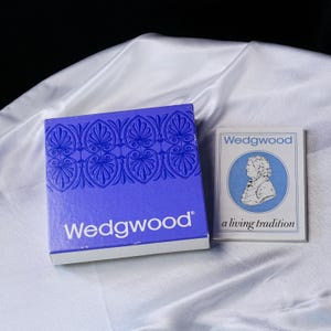 May include: A square blue Wedgwood box featuring a repeating white floral pattern at the top and the white text Wedgwood at the bottom, accompanied by a white rectangular booklet with the text Wedgwood a living tradition and a blue portrait.