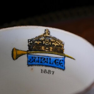 May include: Close-up of a white ceramic bowl with a gold crown and trumpet design. A blue banner reads "JUBILEE" with the year "1887" below. The design features black, gold, and blue accents.