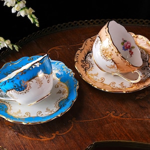 Op de afbeelding: Twee sierlijke kopjes en schotels. E&eacute;n is blauw met gouden randen, de andere is perzik met gouden patronen. Beide staan op een houten dienblad. Witte bloemen op de achtergrond. Voor thee of koffie.