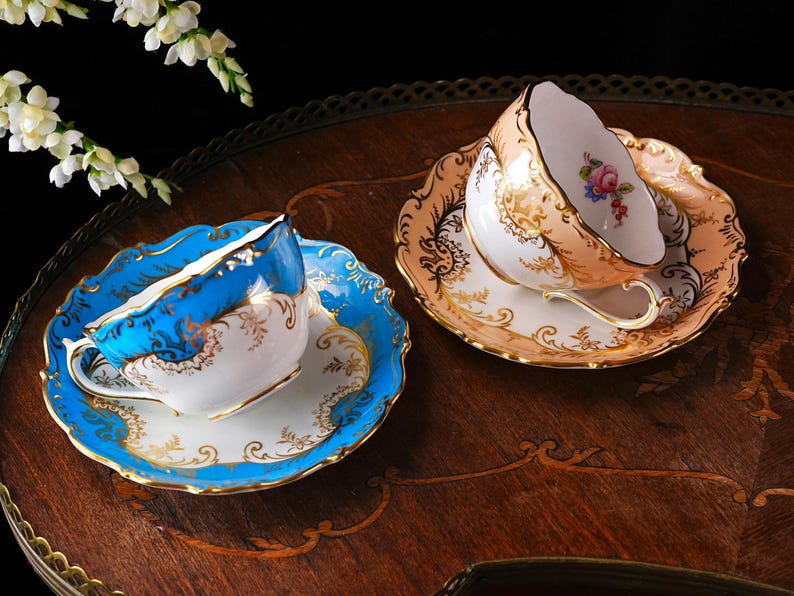 Op de afbeelding: Twee sierlijke kopjes en schotels. E&eacute;n is blauw met gouden randen, de andere is perzik met gouden patronen. Beide staan op een houten dienblad. Witte bloemen op de achtergrond. Voor thee of koffie.