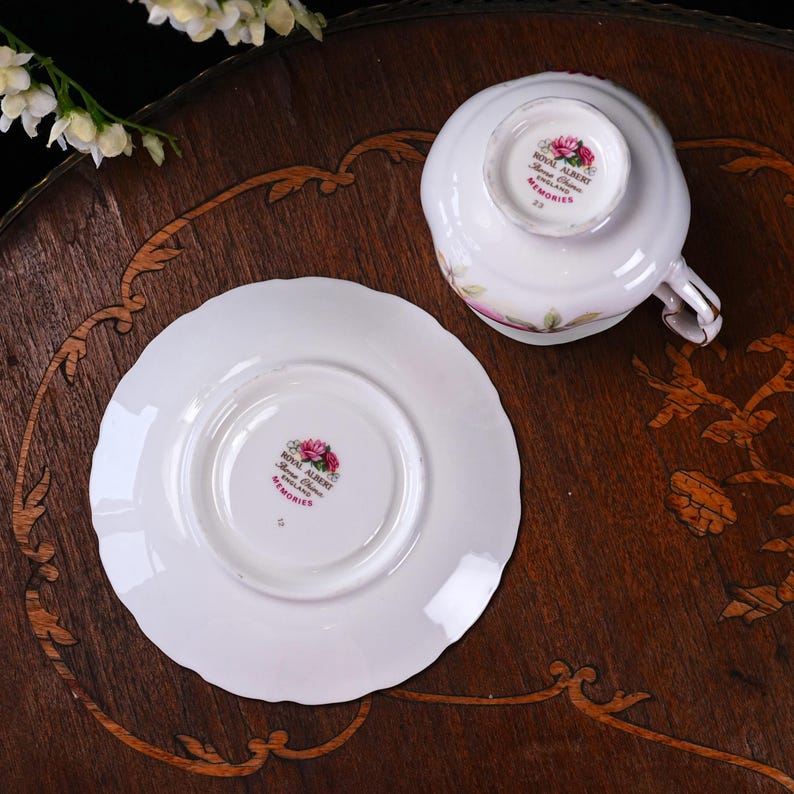 May include: A Royal Albert teacup and saucer set, featuring a floral pattern and gold detailing. The teacup is inverted, displaying the Royal Albert stamp. The saucer has a scalloped edge and the Royal Albert stamp. Set on a dark wooden table.