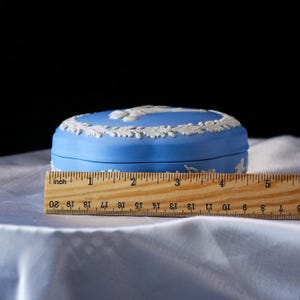 Może przedstawiać: Niebieskie ceramiczne pudełko na drobiazgi z białymi detalami reliefowymi. Pudełko ma owalny kształt i mierzy około 7,6 cm długości. Pokrywa ma dekoracyjne obramowanie i centralny wz&oacute;r. Pudełko stoi na białej powierzchni.
