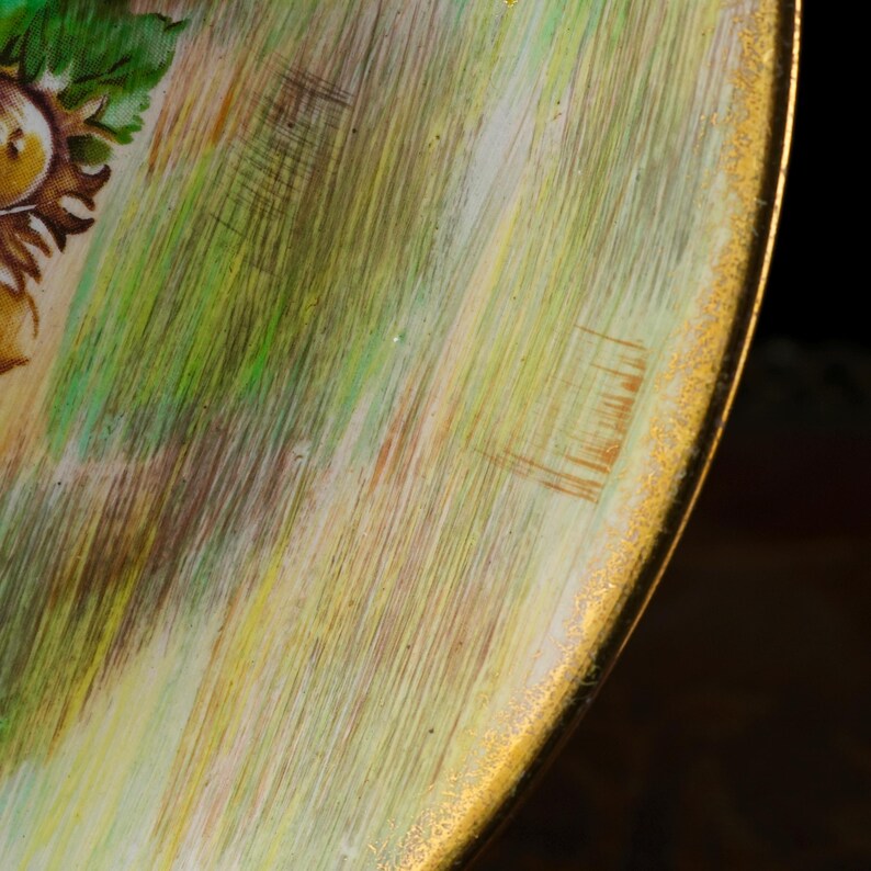 May include: Close-up of a round, painted surface with a gold rim. The surface features a blend of green, yellow, and brown brushstrokes, with a hint of a floral design in the upper left corner. The gold rim adds a touch of elegance.