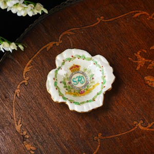Op de afbeelding: Een kleine, witte schaal met gerimpelde rand en gouden rand. De schaal heeft een centraal embleem met een kroon, de letters "SR" en een banner. De rand is versierd met groene bladeren en rode bessen. De schaal staat op een donker houten oppervlak.