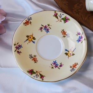 Op de afbeelding: Een cr&egrave;mekleurige schotel met een gouden rand en een wit midden. De schotel is versierd met kleurrijke bloempatronen in tinten roze, blauw, oranje en geel. Het ontwerp is symmetrisch en delicaat.