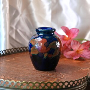May include: A small, bulbous ceramic vase with a deep cobalt blue glaze. The surface features a hand-painted design of rounded orange fruits and green leaves encircling the body. The vase has a short, narrow neck and a smooth, glossy finish.