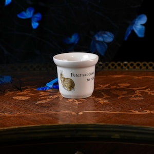 Op de afbeelding: Een kleine witte keramische beker met een illustratie van een bruin konijn en de zwart gedrukte tekst "Peter sat down to rest". De beker heeft een gladde, afgeronde rand en een taps toelopende voet, met een klassiek ontwerp en een citaat.