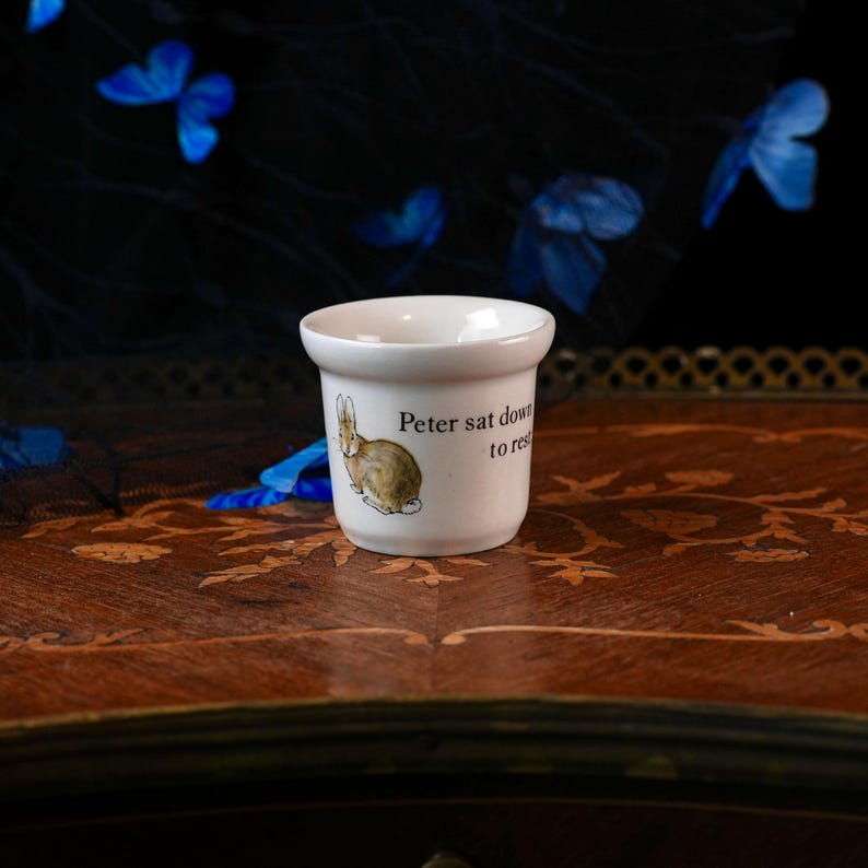 Op de afbeelding: Een kleine witte keramische beker met een illustratie van een bruin konijn en de zwart gedrukte tekst "Peter sat down to rest". De beker heeft een gladde, afgeronde rand en een taps toelopende voet, met een klassiek ontwerp en een citaat.