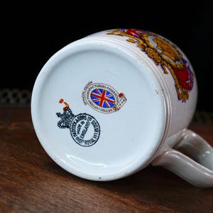 May include: A white ceramic mug with a handle, decorated with red, gold, and blue details. The base of the mug shows the text "MADE IN ENGLAND" and a British Pottery Federation logo.
