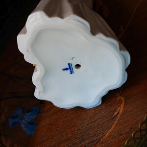 K&ouml;nnte beinhalten: Die Unterseite einer wei&szlig;en Porzellanfigur mit gewelltem Sockel. Der Sockel zeigt ein blau gestempeltes Lladro-Logo, den Text "MADE IN SPAIN", ein kleines kreisf&ouml;rmiges Loch sowie schwache eingepr&auml;gte Markierungen, darunter eine Zahl und ein Symbol.