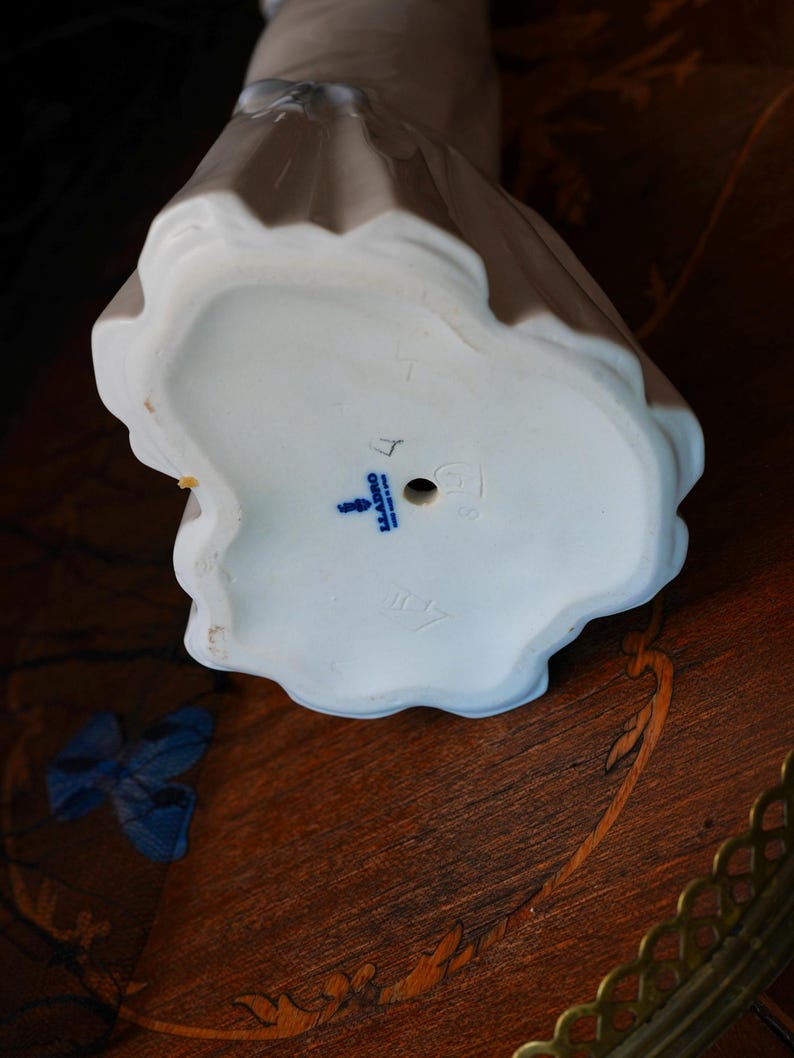 K&ouml;nnte beinhalten: Die Unterseite einer wei&szlig;en Porzellanfigur mit gewelltem Sockel. Der Sockel zeigt ein blau gestempeltes Lladro-Logo, den Text "MADE IN SPAIN", ein kleines kreisf&ouml;rmiges Loch sowie schwache eingepr&auml;gte Markierungen, darunter eine Zahl und ein Symbol.