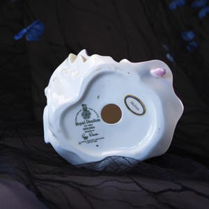 May include: The underside of a white porcelain figurine featuring a circular opening and a Royal Doulton stamp. Text reads Royal Doulton, HN 2467, Melissa, Modelled by Peggy Davies, and Royal Doulton Tableware Limited 1986. A small oval sticker says Melissa.