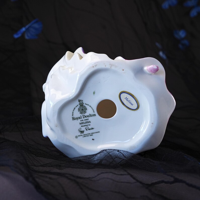 May include: The underside of a white porcelain figurine featuring a circular opening and a Royal Doulton stamp. Text reads Royal Doulton, HN 2467, Melissa, Modelled by Peggy Davies, and Royal Doulton Tableware Limited 1986. A small oval sticker says Melissa.