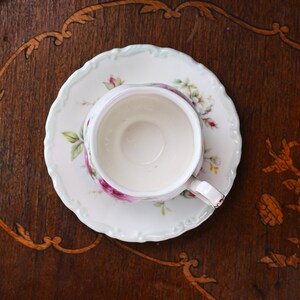 May include: A white teacup and saucer set with a floral pattern, viewed from above. The cup and saucer have a scalloped edge and are adorned with pink roses and green leaves. The set rests on a dark wooden surface.