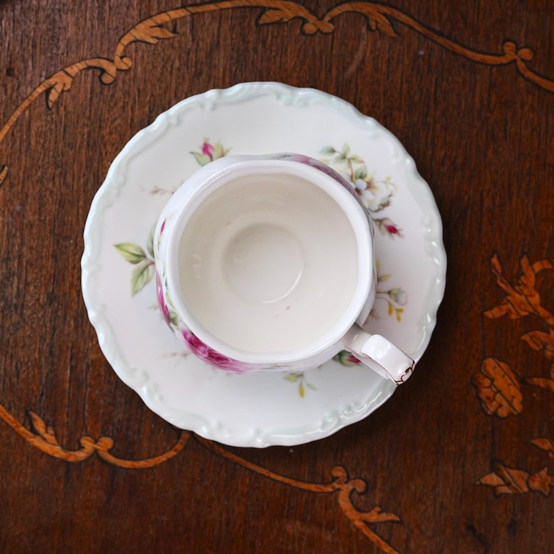 May include: A white teacup and saucer set with a floral pattern, viewed from above. The cup and saucer have a scalloped edge and are adorned with pink roses and green leaves. The set rests on a dark wooden surface.
