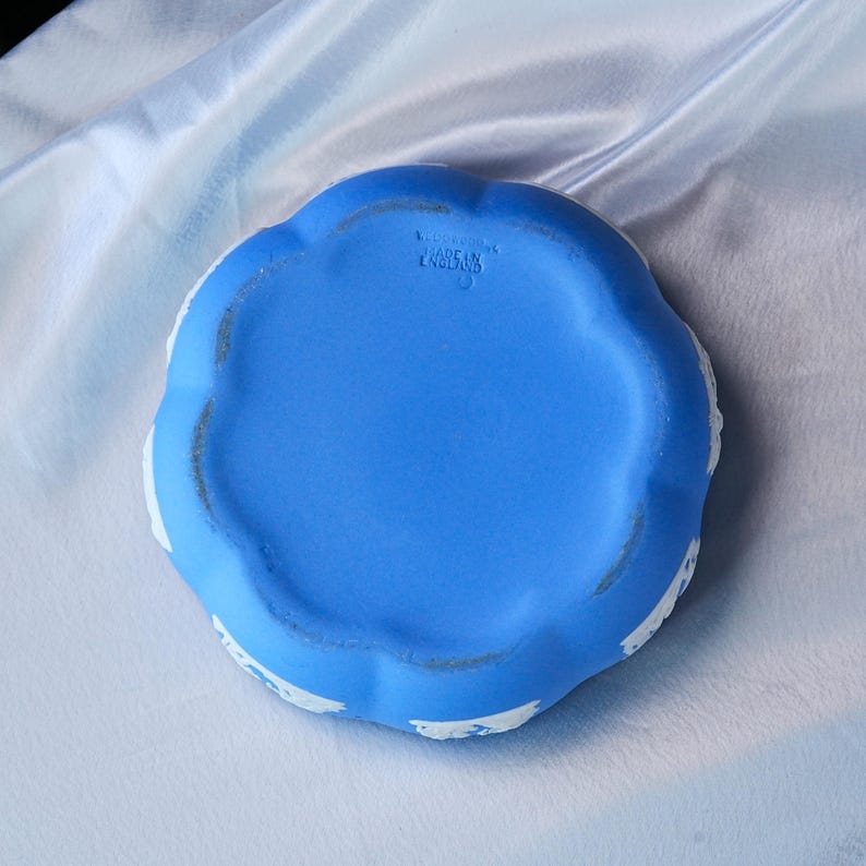 May include: A round, bright blue ceramic dish with a scalloped edge and white relief detailing along the sides. The underside features an embossed stamp that reads WEDGWOOD MADE IN ENGLAND, indicating the origin and manufacturer of the piece.