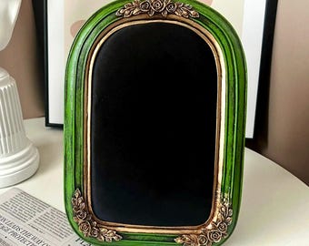 May include: A green and gold framed chalkboard with a black surface. The frame has a decorative design with gold accents and floral details.
