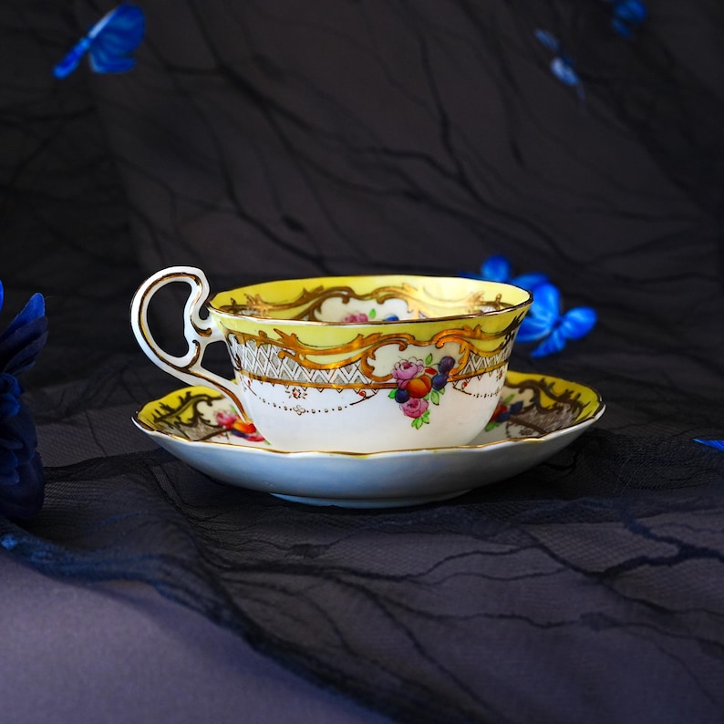 Peut inclure: Un ensemble tasse et soucoupe en porcelaine avec un bord jaune vif orn&eacute; de d&eacute;tails en filigrane dor&eacute;. La surface en c&eacute;ramique blanche est d&eacute;cor&eacute;e d'un petit bouquet color&eacute; de fleurs et de fruits peints, incluant des roses et des baies.