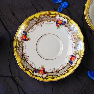 Peut inclure: Une soucoupe en porcelaine ronde &agrave; bord festonn&eacute;, dot&eacute;e d'une bordure jaune vif, d'ornements en filigrane dor&eacute; et d'un motif en treillis. Le centre blanc est d&eacute;cor&eacute; de grappes de p&ecirc;ches color&eacute;es, de baies et de feuilles vertes.