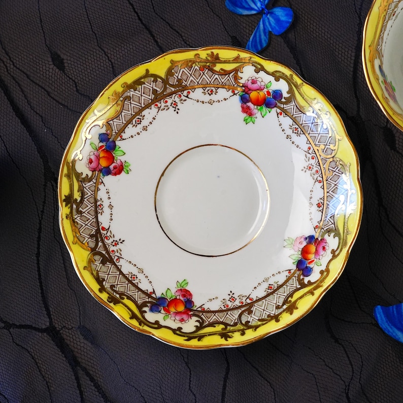 Peut inclure: Une soucoupe en porcelaine ronde &agrave; bord festonn&eacute;, dot&eacute;e d'une bordure jaune vif, d'ornements en filigrane dor&eacute; et d'un motif en treillis. Le centre blanc est d&eacute;cor&eacute; de grappes de p&ecirc;ches color&eacute;es, de baies et de feuilles vertes.