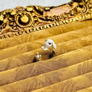 Może przedstawiać: Srebrny pierścionek z detalicznym wzorem królika. Królik jest biały z różowymi oczami i jest ustawiony tak, jakby wychylał się zza czegoś.