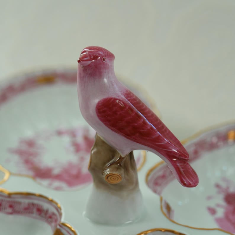 May include: A ceramic bird figurine in shades of pink and red perched on a brown tree branch. The bird has a glossy finish and is set against a backdrop of decorative plates with gold trim and pink floral patterns.