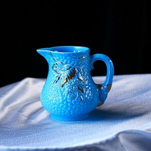 Op de afbeelding: Een felblauw keramieken roomkannetje met een getextureerd, korrelig oppervlak en een opvallend, goudkleurig geschilderd blad- en bessenmotief aan de voorzijde. Het kannetje is voorzien van een gebogen handvat en een kleine schenktuit.
