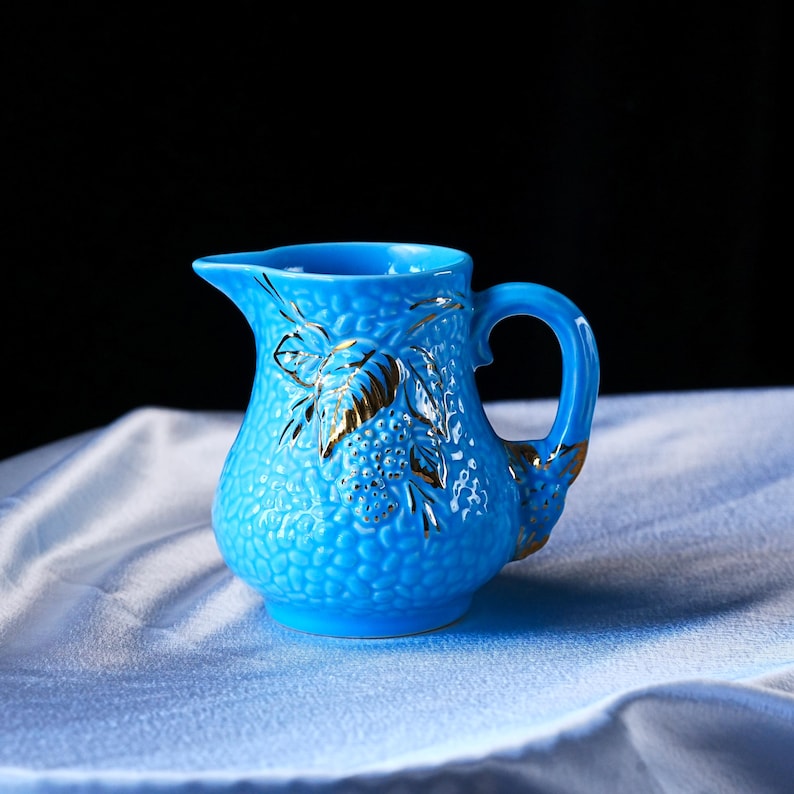 Op de afbeelding: Een felblauw keramieken roomkannetje met een getextureerd, korrelig oppervlak en een opvallend, goudkleurig geschilderd blad- en bessenmotief aan de voorzijde. Het kannetje is voorzien van een gebogen handvat en een kleine schenktuit.