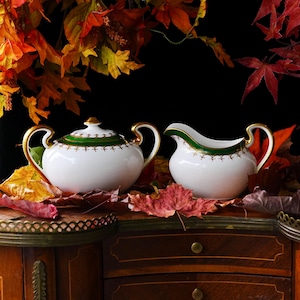 May include: A white porcelain sugar bowl and creamer set with gold handles and green and gold trim. The set is displayed on a wooden surface with autumn leaves.