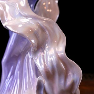 May include: A glossy, pearlescent sculpture with flowing, draped forms. The sculpture has iridescent highlights of lavender, pink, and white. The artwork is set against a dark background, emphasising its curves and textures.