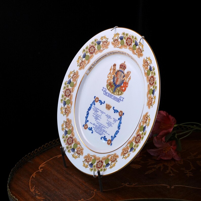 Pode incluir: Um prato comemorativo de porcelana branca com borda dourada, apresentando um bras&atilde;o real central, o texto THE QUEEN MOTHER e uma &aacute;rvore geneal&oacute;gica. A borda &eacute; decorada com um padr&atilde;o repetitivo de cardos e rosas coloridas.