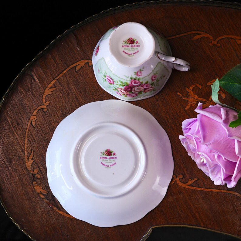 K&ouml;nnte beinhalten: Ein Royal Albert Tee-Set, bestehend aus Tasse und Untertasse. Die wei&szlig;e Porzellantasse und Untertasse haben ein Blumendesign mit rosa Rosen und gr&uuml;nen Akzenten. Das Set wird auf einer Holzoberfl&auml;che pr&auml;sentiert.