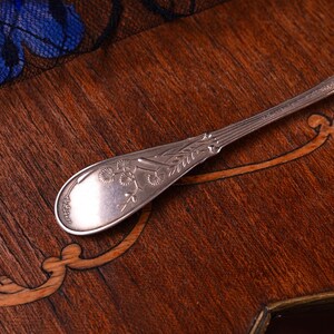 Puede incluir: Un mango de cuchara de metal plateado con un dise&ntilde;o bot&aacute;nico en relieve de peque&ntilde;as hojas en forma de abanico y tallos finos. El mango tiene un extremo redondeado y c&oacute;nico con una serie de estr&iacute;as verticales hacia el cuello.
