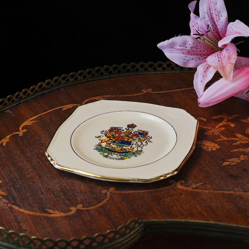 May include: A square-shaped off-white ceramic plate with rounded corners and a gold-trimmed edge. The center features a colorful coat of arms with a lion and unicorn, topped with a crown, and the word CANADA printed in black at the bottom.
