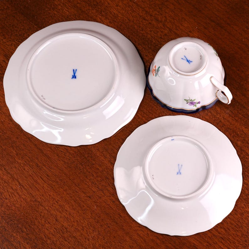 Puede incluir: Una taza de t&eacute; de porcelana blanca y dos platillos con borde festoneado. La taza de t&eacute; tiene un borde azul y dise&ntilde;os florales. Los platillos y la parte inferior de la taza de t&eacute; tienen una marca "X" azul. El juego est&aacute; sobre una superficie de madera.