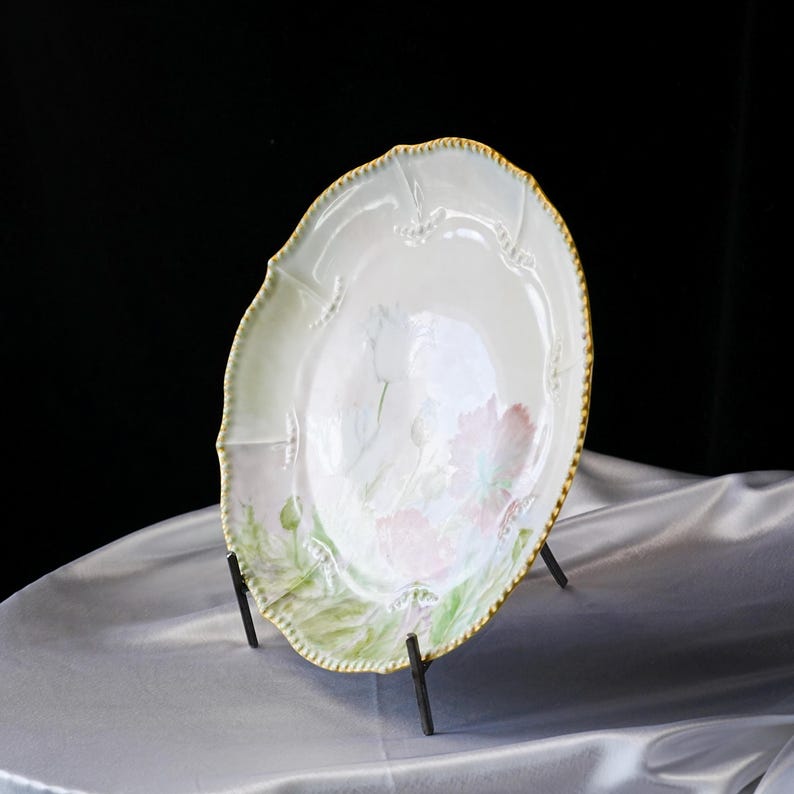 May include: A decorative, antique porcelain plate with a scalloped edge and a gold trim. The plate features a floral design with pink and green accents. The plate is displayed on a black metal stand.