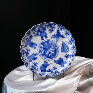 Puede incluir: Un plato de cer&aacute;mica blanca con borde festoneado, con ilustraciones azules. El centro representa un drag&oacute;n, rodeado de figuras y motivos de bamb&uacute;. El plato se exhibe sobre un soporte met&aacute;lico sobre un fondo oscuro.