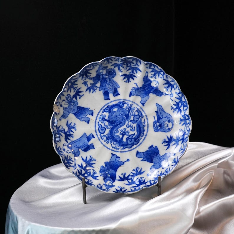 Puede incluir: Un plato de cer&aacute;mica blanca con borde festoneado, con ilustraciones azules. El centro representa un drag&oacute;n, rodeado de figuras y motivos de bamb&uacute;. El plato se exhibe sobre un soporte met&aacute;lico sobre un fondo oscuro.